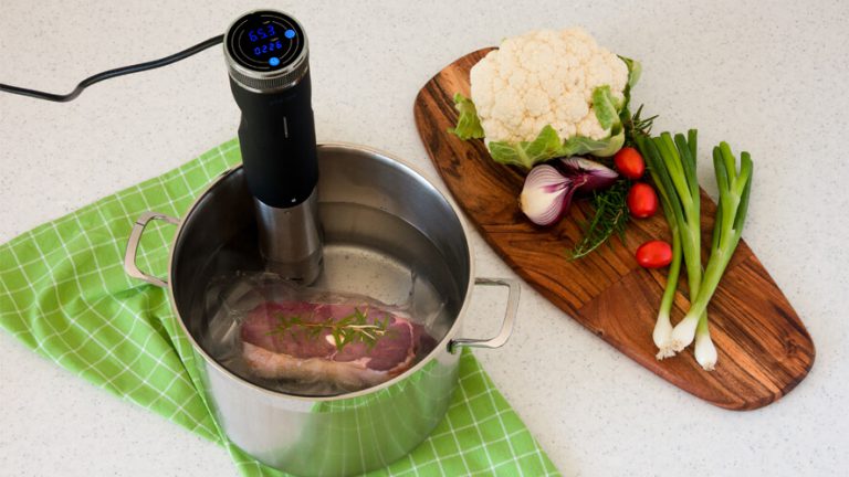 Kuhalnik za počasno kuhanje Status Sous Vide SVC100 pripet na lonec in lesena deska za rezanje z zelenjavo ob strani.